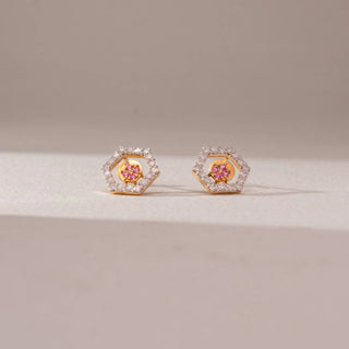 Front view of diamond stud earrings in 9KT yellow gold, featuring hexagonal pavé diamond frames with pink gemstone floral centers, displayed on a beige surface highlighting vibrant contrast and sparkle.
