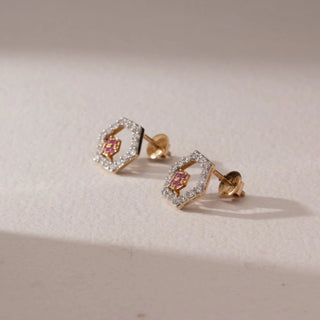 Side view of diamond stud earrings in 9KT yellow gold, featuring hexagonal pavé diamond frames with pink gemstone floral centers and screw backs, set on a beige surface highlighting detail and sparkle.

