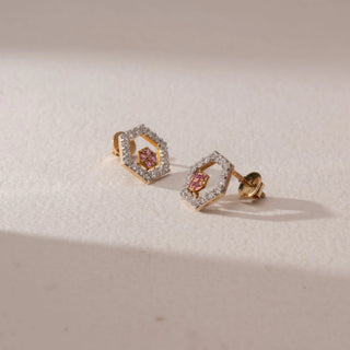 Angled view of diamond stud earrings in 9KT yellow gold, featuring hexagonal pavé diamond frames with pink gemstone floral centers and screw backs, displayed on a beige surface highlighting depth and sparkle.

