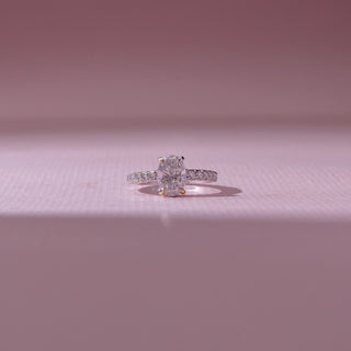 Front view of an oval diamond engagement ring in white gold, featuring a centred oval-cut diamond with pavé-set bands. Captured on a soft pink backdrop, highlighting timeless sparkle and elegance.
