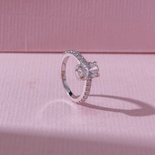 Angled view of a white gold diamond engagement ring, featuring a round-cut centre diamond with pavé-set bands. Captured on a soft pink backdrop, highlighting elegant sparkle and detail.

