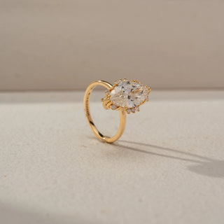 Angled view of a marquise halo engagement ring in 14 KT Yellow Gold, featuring a marquise-cut lab-grown diamond surrounded by a delicate halo of round diamonds, displayed against a beige backdrop.