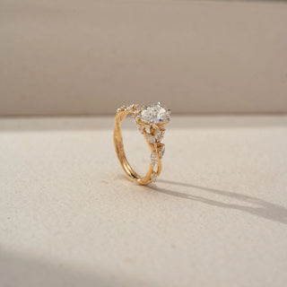 Angled view of a pear-shaped diamond engagement ring in 14 KT Yellow Gold, featuring a pear-cut lab-grown diamond accented by marquise-shaped side diamonds in a nature-inspired band, displayed against a beige backdrop.