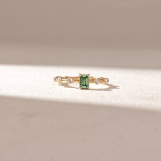 Front view of Emerald-Cut Green Gemstone Ring in 14KT Yellow Gold, featuring a central emerald-cut green stone with round diamond accents on the band. Set on a soft beige surface, it reflects refined elegance.
