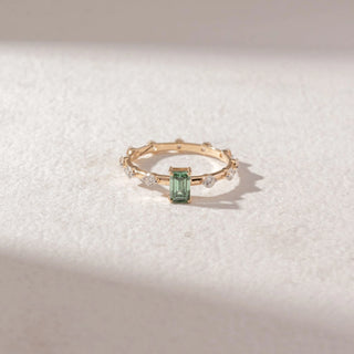 Top view of Emerald-Cut Green Gemstone Ring in 14KT Yellow Gold, featuring a central emerald-cut green stone with round diamond accents along the band. Set on a soft beige surface, it reflects delicate elegance.
