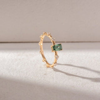 Angled view of Emerald-Cut Green Gemstone Ring in 14KT Yellow Gold, featuring a central emerald-cut green stone with spaced round diamond accents. Set on a soft beige surface, it reflects delicate brilliance.
