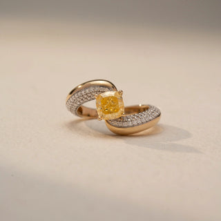 Front view of a bypass diamond ring in 14 KT Yellow Gold, featuring a cushion-cut yellow center diamond embraced by pavé-set diamond bands, displayed against a beige backdrop.