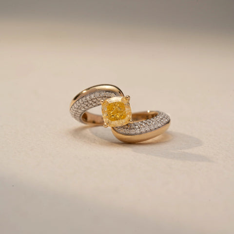 Front view of a bypass diamond ring in 14 KT Yellow Gold, featuring a cushion-cut yellow center diamond embraced by pavé-set diamond bands, displayed against a beige backdrop.