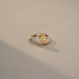 Side view of a bypass diamond ring in 14 KT Yellow Gold, featuring a cushion-cut yellow center diamond embraced by pavé-set diamond bands, displayed against a beige backdrop.