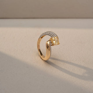 Angled view of a bypass diamond ring in 14 KT Yellow Gold, featuring a cushion-cut yellow center diamond embraced by pavé-set diamond bands, displayed against a beige backdrop.