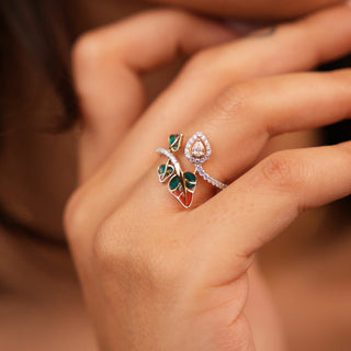 A front view of a female ring of size 13 laying gently on a finger, crafted in 14 KT Gold and 0.36 carat Lab Grown diamond and the weight of the product is 2.35 grams. It features a leafy pattern, with emerald-coloured accents and is surrounded by smaller diamonds arranged in a curved, floral-inspired pattern. The band is made of yellow gold with decorative metalwork.