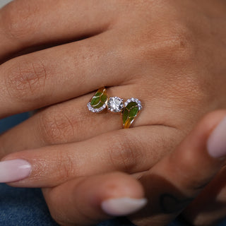 A close-up top view of a female ring of size 13 modelled on a hand, featuring a central round brilliant lab-grown diamond, 0.6 carats with a solitaire of 0.50 carats, flanked by two marquise-shaped green gemstones, total weight is 2.15 grams. The setting is crafted from 14 KT yellow gold, creating a visually striking and elegant piece of jewellery.