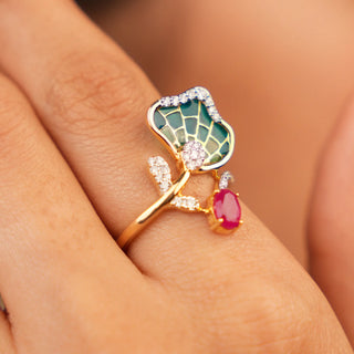 A top view of the nature-inspired female ring of size 12  modelled on a hand, featuring a pink gemstone accented by smaller round diamonds. The centrepiece is a vibrant ruby complemented by an ornate green gemstone motif, arranged in a floral or leaf design. The lab-grown diamond ring band is crafted from 14 KT yellow gold metalwork and 0.26 carats and the weight of the product is 3.38 grams