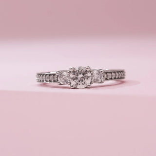 Front view of Reia’s 14 KT White Gold Threefold Promise Solitaire Diamond Ring, showing three round diamonds and a pavé band. Set against pink, it highlights a symmetrical, timeless layout.