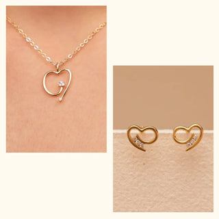 Open-heart motif pendant necklace paired with matching stud earrings, shown in worn and product views, featuring subtle diamond accents highlighted under soft, warm lighting.