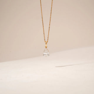 Front view of a pear-shaped diamond pendant in 14KT Yellow Gold, suspended from a delicate gold chain against a beige backdrop.