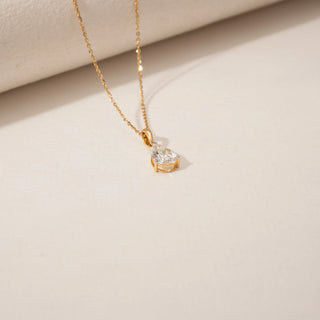 Top view of a pear-shaped diamond pendant in 14KT Yellow Gold, suspended from a delicate gold chain against a beige backdrop.