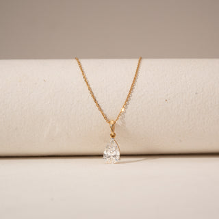Close-up view of a pear-shaped diamond pendant in 14KT Yellow Gold, suspended from a delicate gold chain against a beige backdrop.