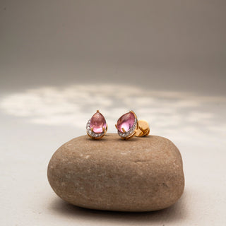 Angled view of pear-shaped pink gemstone stud earrings in 14 KT Yellow Gold, featuring soft pink center stones framed with pavé diamond accents, displayed on a textured stone surface.