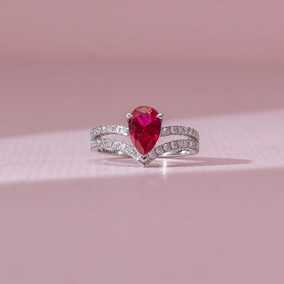 Front view of red diamond ring in white gold, featuring a pear-shaped vibrant red centre stone with a split diamond-studded band of round lab-grown diamonds. Set against a soft pink backdrop, it reflects bold elegance.