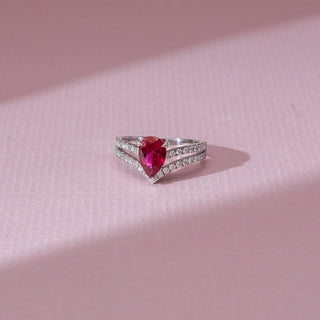 Top view of red diamond ring in white gold, featuring a pear-shaped vibrant red centre stone with a split diamond-studded band of round lab-grown diamonds. Set on a soft pink surface, it highlights bold brilliance.