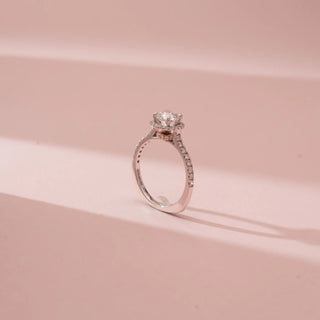 Angled view of Reia’s 14 KT White Gold Classic Halo Solitaire Diamond Ring. It highlights the diamond-set band, shimmering halo, and six-prong set center diamond on a soft pink backdrop.