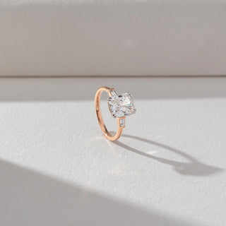 Angled view of Radiant Solitaire Diamond Ring in 14KT Rose Gold, featuring a centred radiant-cut lab grown diamond with baguette side accents. Set on a beige backdrop, the design highlights depth and brilliance.
