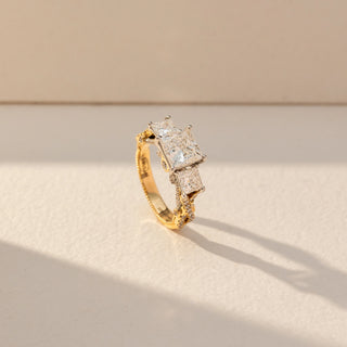 Side view of Princess Three Stone Diamond Ring in 14KT Yellow Gold, featuring a centred princess-cut lab grown diamond with square side stones. Set on a beige backdrop, the design highlights depth and brilliance.
