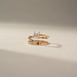 Front view of an emerald-cut bypass ring in 14 KT Rose Gold, featuring an emerald-cut lab-grown diamond and channel-set baguette diamonds on a sleek open wrap design, displayed against a beige backdrop.