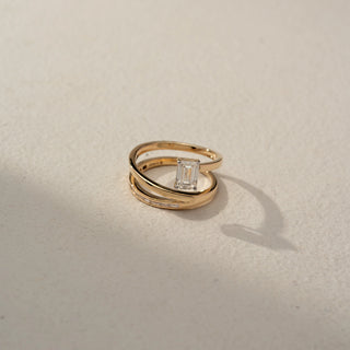 Top view of an emerald-cut bypass ring in 14 KT Rose Gold, featuring an emerald-cut lab-grown diamond and channel-set baguette diamonds on a sleek open wrap design, displayed against a beige backdrop.