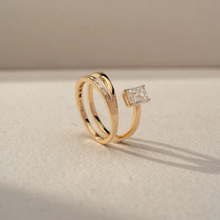 Side view of an emerald-cut bypass ring in 14 KT Rose Gold, featuring an emerald-cut lab-grown diamond and channel-set baguette diamonds on a sleek open wrap design, displayed against a beige backdrop.