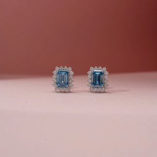 Front view of blue  diamond earrings in 14KT White Gold, featuring emerald-cut diamond and a diamond halo against a pink backdrop.