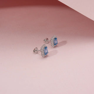 Side view of blue diamond earrings in 14KT White Gold, featuring emerald-cut diamond and a diamond halo against a pink backdrop.