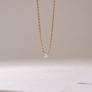 Front view of a delicate yellow gold pendant necklace, featuring a rectangular solitaire diamond suspended on a fine chain. Captured against a soft beige backdrop, the design reflects minimal elegance and sparkle.