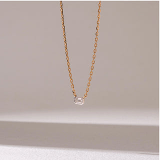 Angled view of a delicate yellow gold pendant necklace, featuring a rectangular solitaire diamond suspended on a fine chain. Captured against a soft beige backdrop, the design reflects minimal elegance and sparkle.