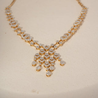 Front view of a diamond necklace in 14 KT Yellow Gold, featuring interlinked pavé-set oval motifs and a cascading diamond cluster centerpiece with pear-shaped drops against a soft beige backdrop.

