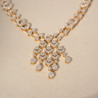 Close up view of a diamond necklace in 14 KT Yellow Gold, featuring interlinked pavé-set oval motifs and a cascading diamond cluster centerpiece with pear-shaped drops against a soft beige backdrop.
