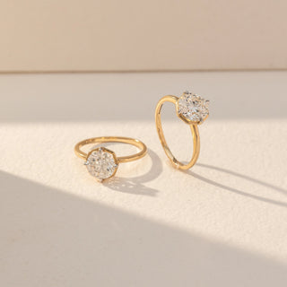 Angled view of Round Solitaire Diamond Rings in 14KT Yellow Gold, featuring round lab grown diamonds in prong settings. Set on a beige backdrop, the design highlights classic elegance and brilliance.
