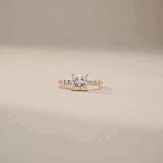 Front view of a princess-cut solitaire engagement ring in 14 KT Yellow Gold, featuring a princess-cut lab-grown diamond accented by heart-shaped side diamonds along the band, displayed against a beige backdrop.