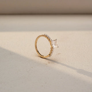 Angled view of a princess-cut solitaire engagement ring in 14 KT Yellow Gold, featuring a princess-cut lab-grown diamond accented by heart-shaped side diamonds along the band, displayed against a beige backdrop.