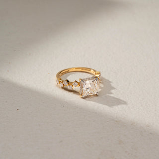 Side view of a princess-cut solitaire engagement ring in 14 KT Yellow Gold, featuring a princess-cut lab-grown diamond accented by round side diamonds along the band, displayed against a beige backdrop.