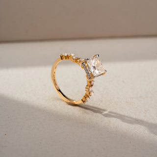 Angled view of a princess-cut solitaire engagement ring in 14 KT Yellow Gold, featuring a princess-cut lab-grown diamond accented by round side diamonds along the band, displayed against a beige backdrop.