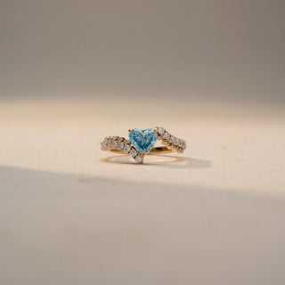 Front view of a heart-cut blue gemstone ring in 14 KT Yellow Gold, featuring a heart-shaped blue center stone embraced by a diamond-accented bypass band, displayed against a beige backdrop.