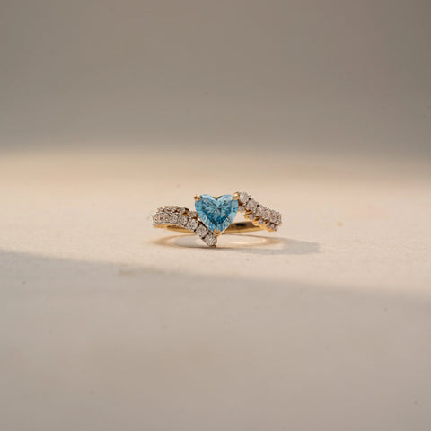 Front view of a heart-cut blue gemstone ring in 14 KT Yellow Gold, featuring a heart-shaped blue center stone embraced by a diamond-accented bypass band, displayed against a beige backdrop.