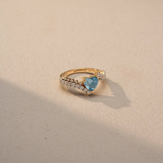 Side view of a heart-cut blue gemstone ring in 14 KT Yellow Gold, featuring a heart-shaped blue center stone embraced by a diamond-accented bypass band, displayed against a beige backdrop.