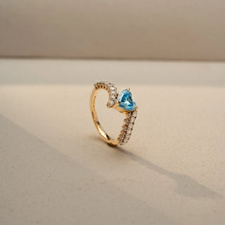 Angled view of a heart-cut blue gemstone ring in 14 KT Yellow Gold, featuring a heart-shaped blue center stone embraced by a diamond-accented bypass band, displayed against a beige backdrop.