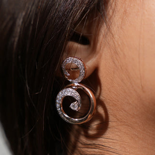 A close-up side view of an female earring modelled on an ear featuring a swirl design crafted in 14 KT rose gold. Each earring has a round brilliant-cut diamond at the centre, surrounded by a spiral of smaller round diamonds set into the rose gold metal work weighing 13.41 grams secured through a bombay screw.  The total carat weight of the lab-grown diamonds used in this intricate earring pair is 1.103 carats, creating a sparkling and eye-catching look.