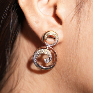 A close-up side view of an earring modelled on an ear featuring a swirl design crafted in 14 KT rose gold. Each earring has a round brilliant-cut diamond at the centre, surrounded by a spiral of smaller round diamonds set into the rose gold metal work weighing 13.41 grams secured through a bombay screw. The total carat weight of the lab-grown diamonds used in this intricate earring pair is 1.103 carats, creating a sparkling and eye-catching look.