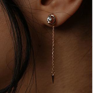 A close-up side view of an female earring modelled on an ear featuring a 14 KT rose gold setting with a round diamond stud on the earlobe connected by a chain to a dangling spike pendant weighing 2.39 grams secured through a bombay screw. The total carat weight of the lab-grown diamonds is 0.24 carats, showcasing an elegant and trendy style.