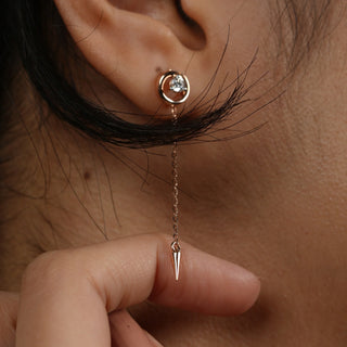 A close-up front view of an female earring modelled on an ear featuring a 14 KT rose gold setting with a round diamond stud on the earlobe connected by a chain to a dangling spike pendant weighing 2.39 grams secured through a bombay screw. The total carat weight of the lab-grown diamonds is 0.24 carats, showcasing an elegant and trendy style.